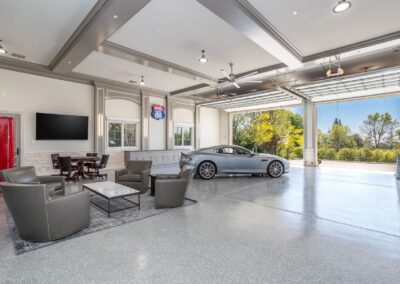 Spacious modern garage with a silver car, lounge area with chairs and table, large TV, and open door revealing a view of trees outside.