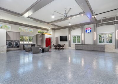 Spacious room with high ceilings, featuring a seating area, red refrigerator, wall-mounted TV, and laundry machines. Retro sign and large windows are also visible.