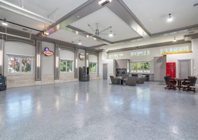 A large, empty room with a glossy floor, two sofas, a dining table, and various neon signs on the walls. A washing machine and refrigerator are in the corner. Large windows let in natural light.