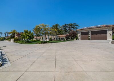 Spacious driveway leading to a large, modern house with a well-maintained lawn, palm trees, and a three-car garage under a clear blue sky.