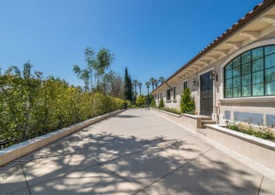 A wide driveway leads to a building with arched windows and a tiled roof, bordered by greenery and palm trees under a clear blue sky.