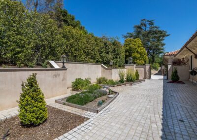 Paved courtyard with raised garden beds, small shrubs, and wall-mounted lanterns, bordered by a high wall and greenery under a clear blue sky.