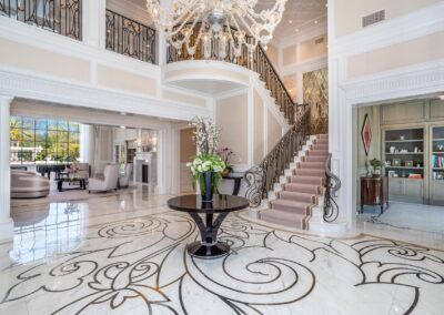 Opulent foyer with a grand staircase, intricate marble floor design, chandelier, and a round table with flowers. Open view to a luxurious living area with large windows and furnishings.