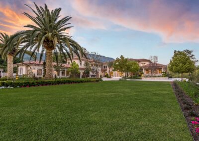 A large house with a manicured lawn, tall palm trees, and a mountainous backdrop under a colorful sunset sky.