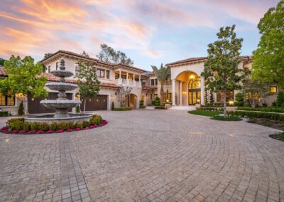 Luxurious mansion with Mediterranean architecture, featuring a circular driveway, central fountain, and manicured gardens at sunset.