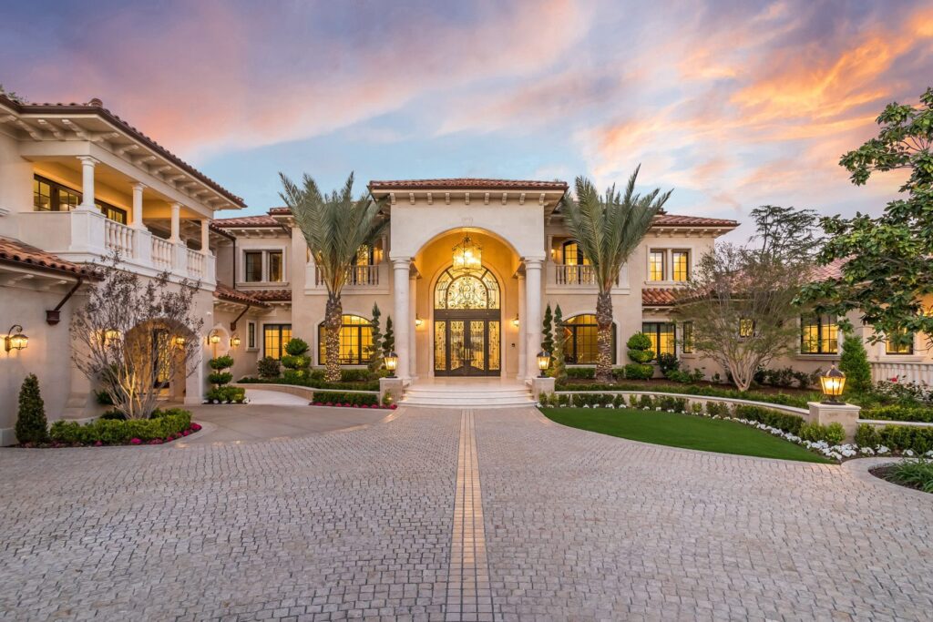 A luxurious mansion with large arched entryway, surrounded by palm trees and a circular driveway under a colorful sunset sky.