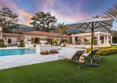 Luxurious villa with a pool, sun loungers, and a large umbrella, surrounded by manicured gardens and trees under a colorful sunset sky.