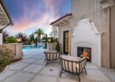 Outdoor patio with two cushioned chairs facing a fireplace. In the background, a pool and palm tree are visible under a colorful sky.