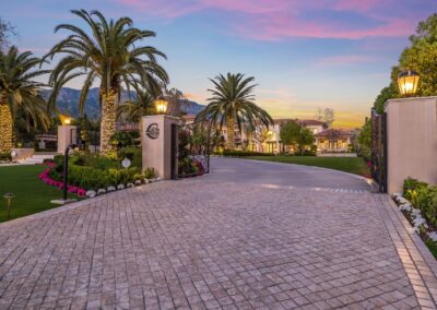 Gated entrance to a luxurious estate with palm trees, a cobblestone driveway, and well-manicured lawns during sunset.