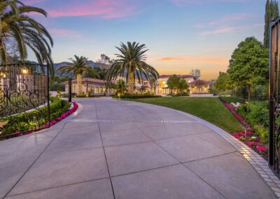 A paved driveway leads to a large house surrounded by manicured lawns, palm trees, and flowers under a colorful sunset sky.