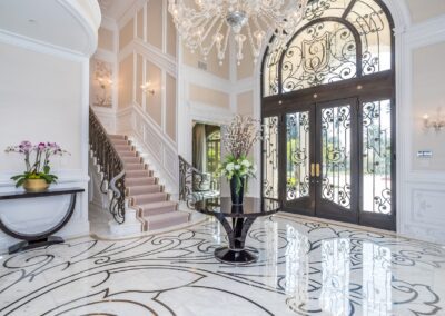 Luxurious foyer with marble floor, grand staircase, ornate chandelier, and large decorative glass doors. A table with flower arrangements is centrally placed.