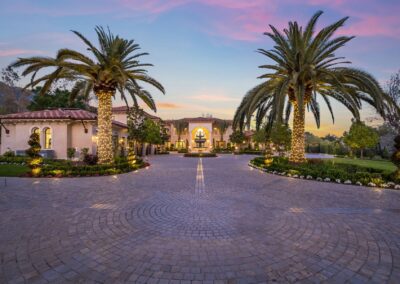 Large estate with a circular driveway, palm trees wrapped in lights, manicured gardens, and a grand entrance. A colorful sunset is in the background.