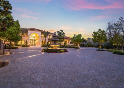 A large, elegant mansion with a grand entrance, circular driveway, and central fountain, surrounded by trees at sunset.