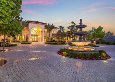 Large mansion with illuminated entrance, circular driveway, and tiered fountain surrounded by flowers, set against a sunset sky.