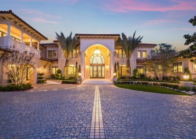 A grand mansion with a large arched entrance, palm trees, and a circular cobblestone driveway, set against a sunset sky.