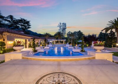 Luxurious estate with a large illuminated pool, surrounded by elegant architectural features, palm trees, and a landscaped garden at dusk.