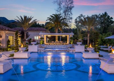 Luxurious outdoor pool area with fountains, surrounded by palm trees and landscaping. A pavilion with seating is visible in the background, all set during twilight.