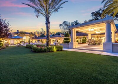 Spacious backyard with well-manicured lawn, palm trees, and a covered outdoor seating area next to a large, illuminated house at sunset.