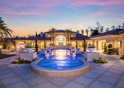 Luxury mansion with elegant architecture, large windows, and a central fountain. Lush greenery surrounds the property, set against a vibrant sunset sky.