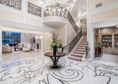 Elegant foyer with a spiral staircase, chandelier, marble floors with intricate designs, a round table with floral arrangement, and a sitting area with a fireplace on the left.