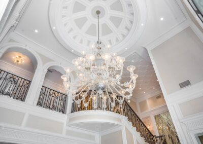 Ornate chandelier hanging from a domed ceiling in a grand staircase area with intricate iron railings and large windows.