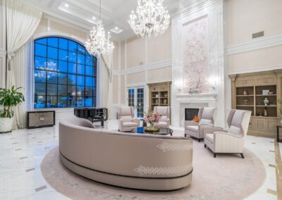 Elegant living room with large window, chandeliers, and cream-colored furniture arranged on a circular rug. High ceilings and a fireplace with intricate wall art create a luxurious atmosphere.