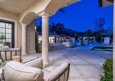 Patio view of a luxurious outdoor area at dusk, featuring columns, cushioned patio chairs, a pool, and surrounding greenery.