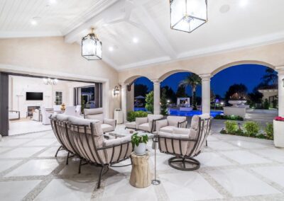 Luxurious outdoor patio with elegant seating, chandeliers, and arches overlooking a pool and garden. The space is well-lit, combining indoor and outdoor elements seamlessly.