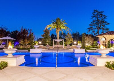 Luxurious pool area with a central palm tree, fountains, and lounge chairs under canopies. Surrounded by greenery, the scene is set against a clear, vibrant blue evening sky.