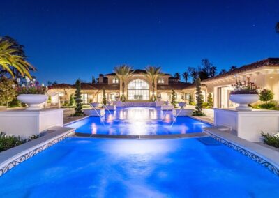 Luxurious mansion with a grand, illuminated pool in the foreground, surrounded by palm trees and elegant architecture, set against a clear evening sky.