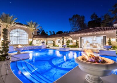 Luxury mansion with a large swimming pool, surrounded by lit fire bowls and palm trees, at dusk. The house features an arched window and open doors to a well-lit interior.