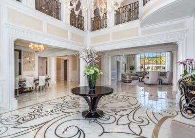 Luxurious foyer with intricate marble flooring, central black table with floral arrangement, and elegant double-height ceiling. Dining and living areas visible beyond.