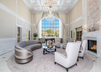 Luxurious living room with a large window, elegant chandelier, curved sofa, armchairs, and a round coffee table. Marble fireplace on the right and decorative floral accents throughout.