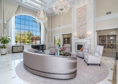 Spacious living room with high ceilings features a large, curved gray sofa, elegant armchairs, a chandelier, and a tall window overlooking a garden.