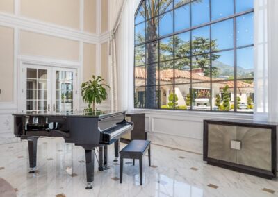 A grand piano with a bench sits on a marble floor in a spacious room with a large arched window and chandelier.