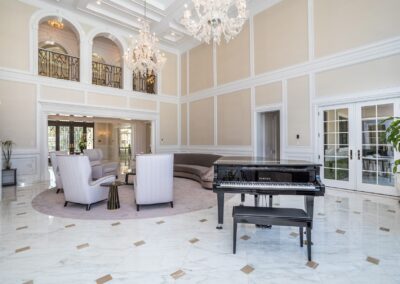 A spacious, elegantly decorated living room with marble flooring, a grand piano, high ceilings, chandeliers, and white furniture arranged around a circular rug.