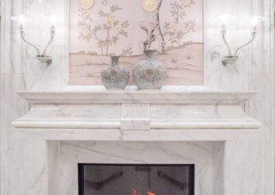 A marble fireplace with a lit fire, flanked by two patterned vases. Elegant wall sconces and decorative floral artwork are displayed above.