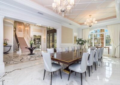 Elegant dining room with a long table and white chairs, marble floors, chandelier lighting, and a view of a grand staircase through an open doorway.