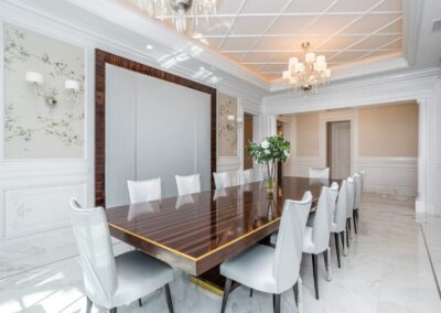 Elegant dining room with a long, glossy table surrounded by white chairs. Floral wallpaper and chandeliers enhance the sophisticated atmosphere.