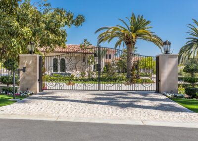 A large gated driveway leads to a house with a tiled roof, surrounded by palm trees and manicured gardens.