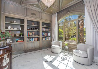 A luxurious library with built-in bookshelves, two white armchairs, a large arched window, and a chandelier, all set in a room with marble flooring.