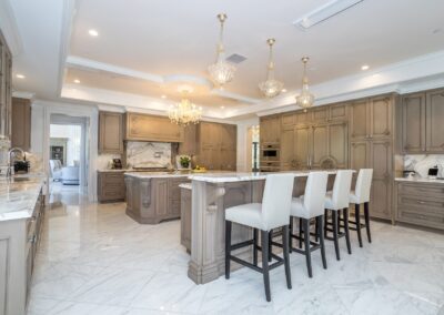 Spacious kitchen with marble floors, large island with white chairs, wooden cabinets, and chandeliers.