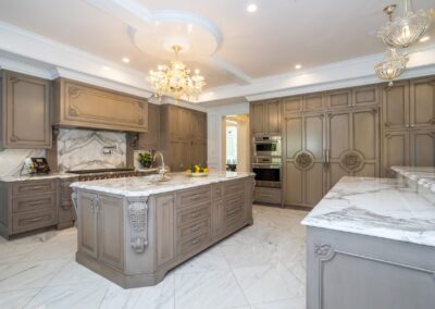 Luxurious kitchen with marble countertops, large island, ornate wood cabinetry, chandeliers, double ovens, and white tile flooring.