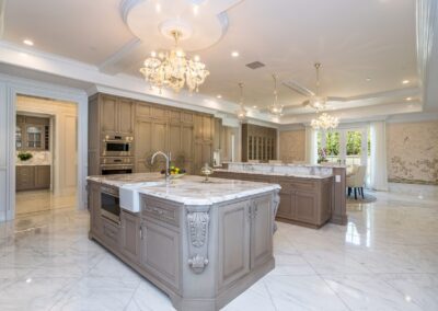 Spacious kitchen with marble countertops, chandeliers, and ornate wood cabinetry. Two central islands with sinks and integrated appliances.