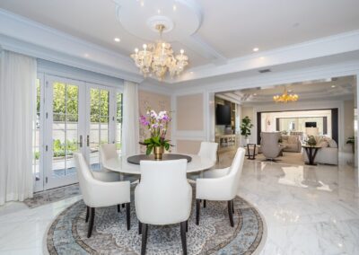 Luxurious dining room with a round table, white chairs, marble flooring, elegant chandelier, and large windows leading to an outdoor view.