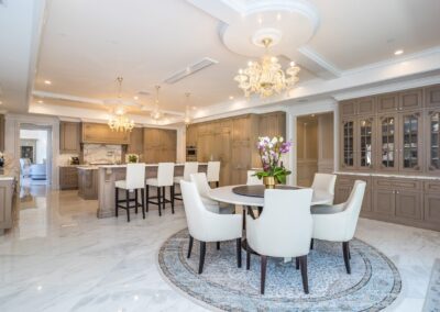 Spacious luxury kitchen with marble floors, two chandeliers, beige cabinetry, a central island with bar stools, and a round dining table with white chairs on a patterned rug.