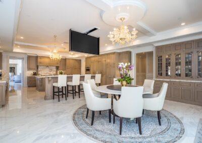 Spacious modern kitchen with a round dining table and chairs on a patterned rug, chandeliers, and a flat-screen TV mounted above the dining area.