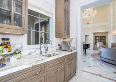 Luxurious kitchen with marble countertops, wooden cabinets, and a window. Nearby, a spacious room features a round sofa, chairs, and a grand piano under a chandelier.