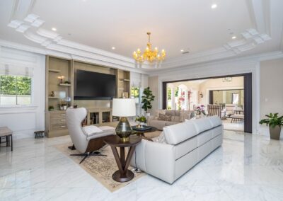 Luxurious living room with white marble floors, a white sofa, a chandelier, and a flat-screen TV. Large windows and sliding doors open to a sunlit patio.