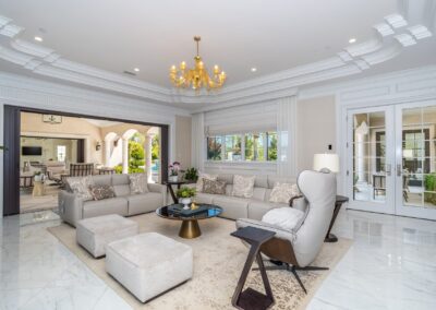 Spacious luxurious living room with white sofas, armchairs, a central coffee table, a golden chandelier, and marble flooring. Large windows and doors offer views of a connected room and greenery.